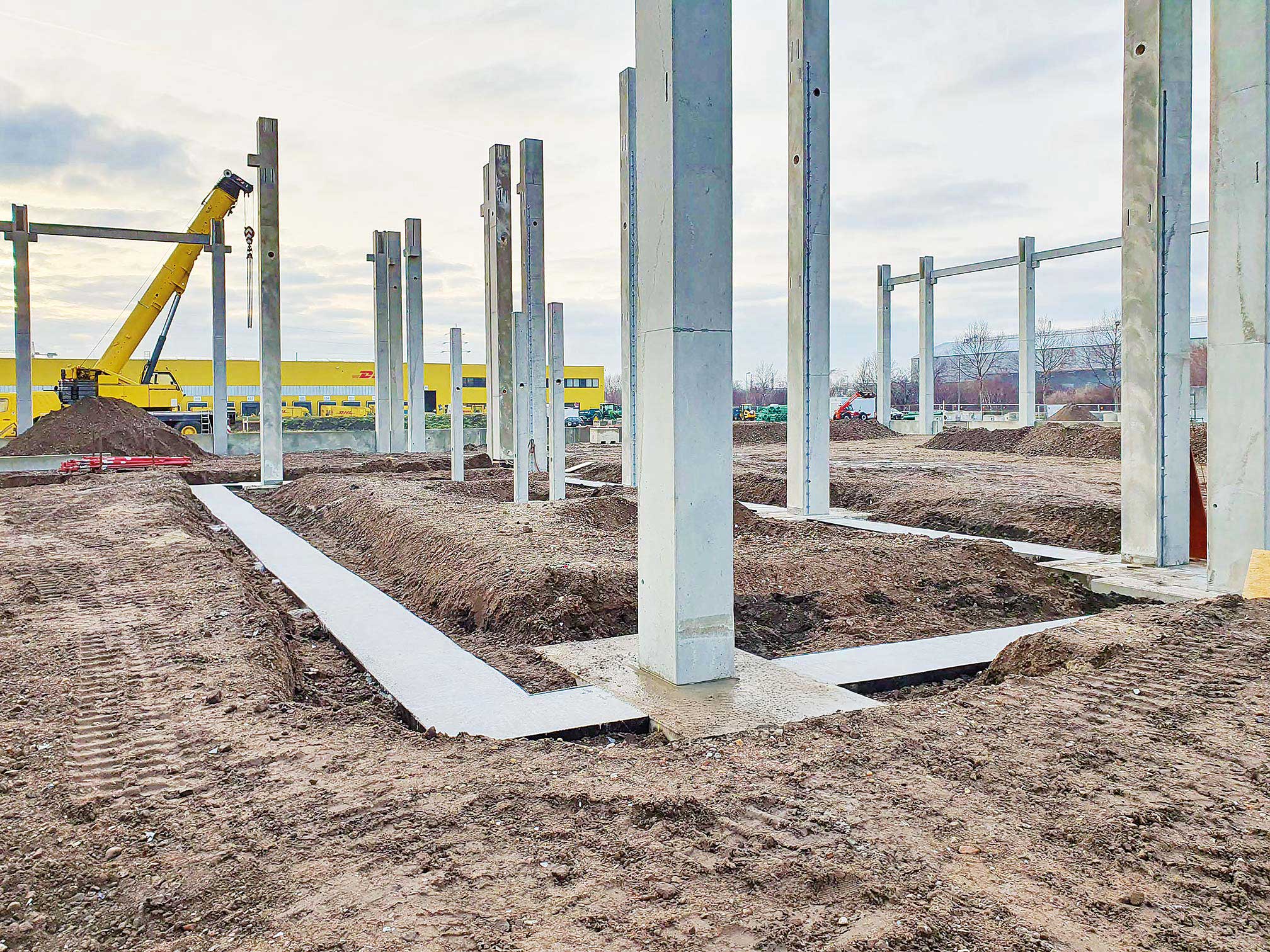 Spezialist für Bau und Anlagenbau Baustelle mit Betonpfeilern und einem gelben Kran vor einem gelben Gebäude bei bewölktem Himmel.