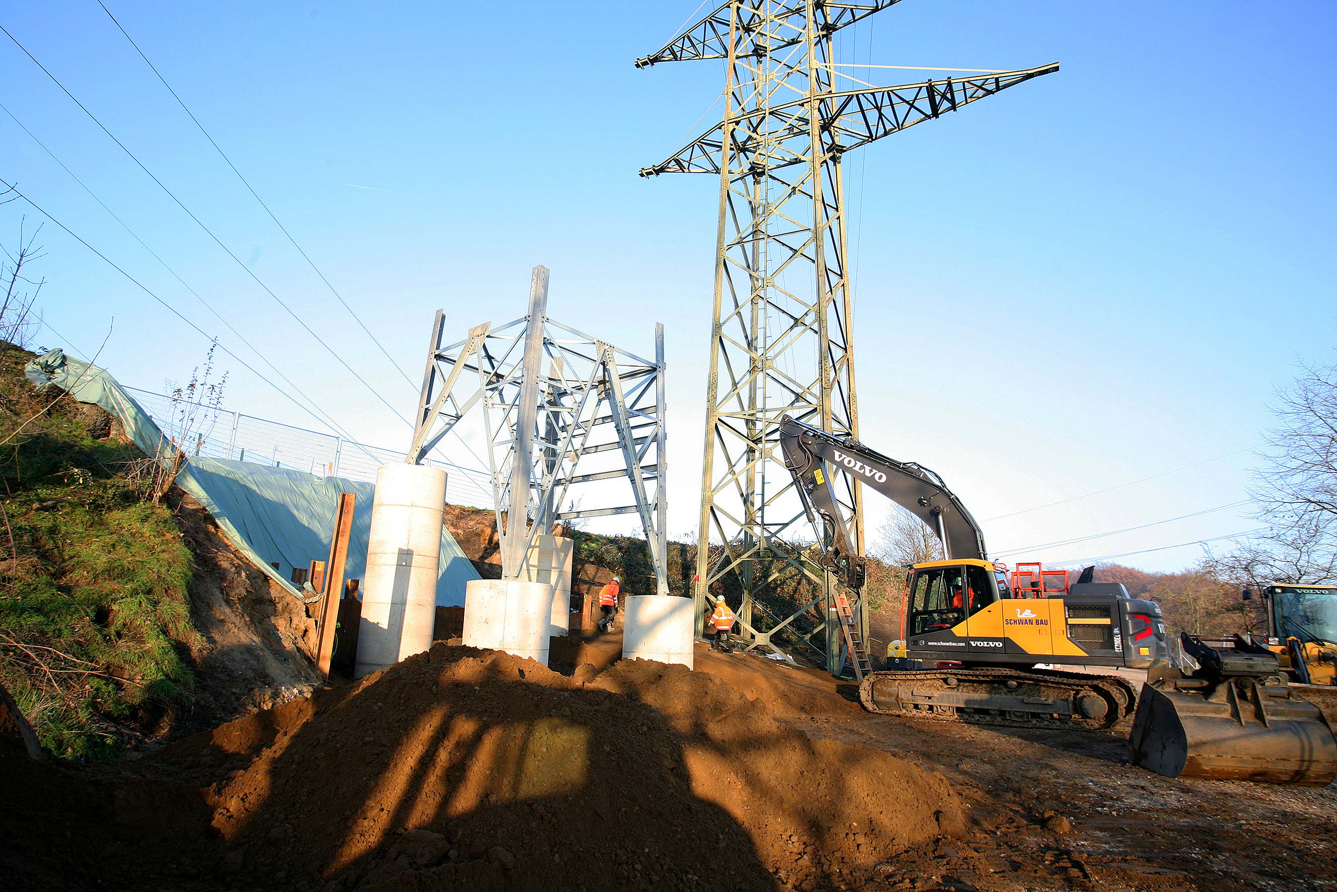 Werde Teil unseres Teams Baustelle mit Volvo-Bagger, Bauarbeitern und Stahlkonstruktion unter Hochspannungsmasten bei klarem Himmel.